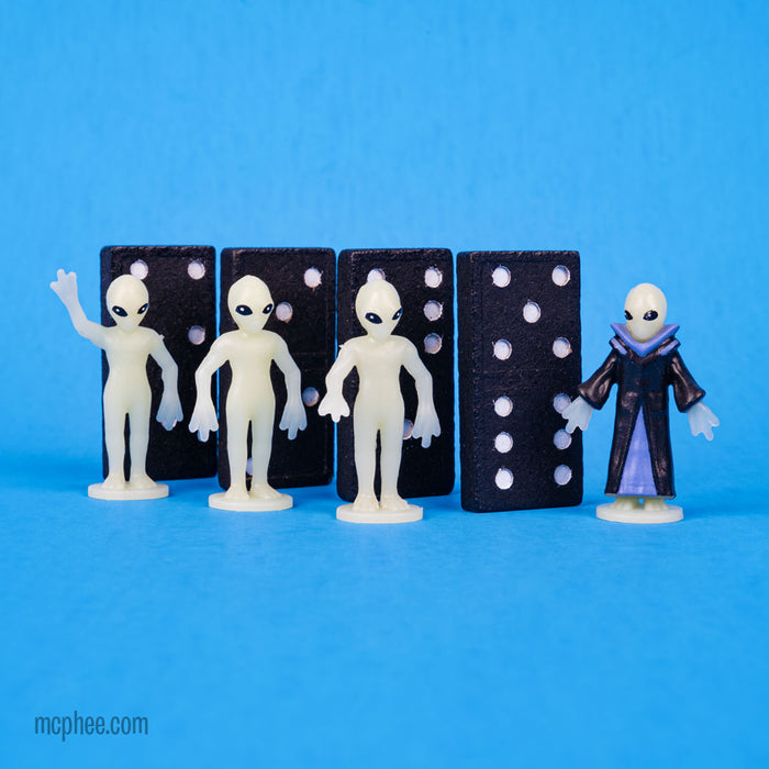 The Itty Bitty Aliens are set against a blue background with dominoes, their distinctive glow-in-the-dark features enhancing their appearance. Among them, one wears a striking black cape, bringing an enigmatic flair to the display.