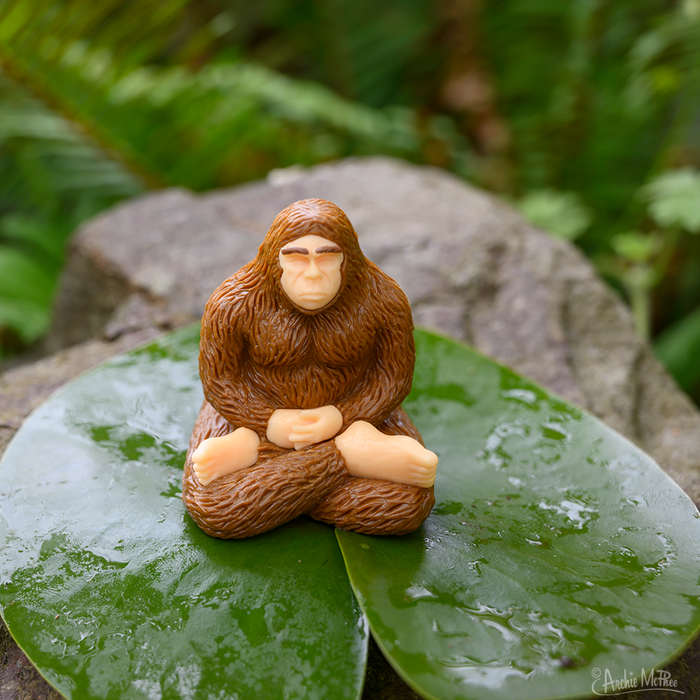 The Meditating Bigfoot collectible figure sits cross-legged on wet green leaves, embodying a serene mindfulness in its meditative pose.