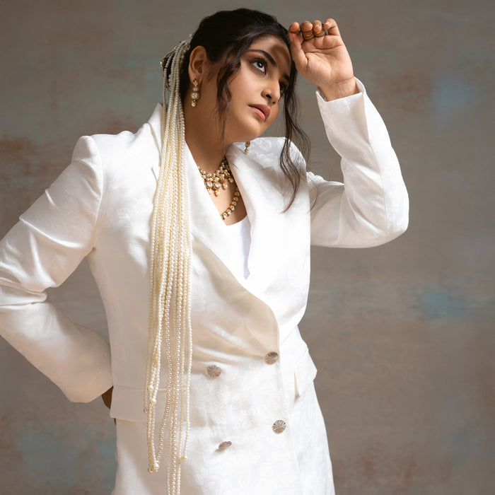 Woman in a white outfit with long pearl necklace against a neutral background