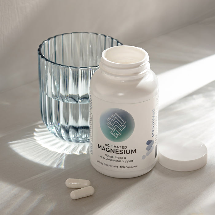Infiniwell Activated Magnesium supplement bottle with open cap, two capsules and glass of water on light surface