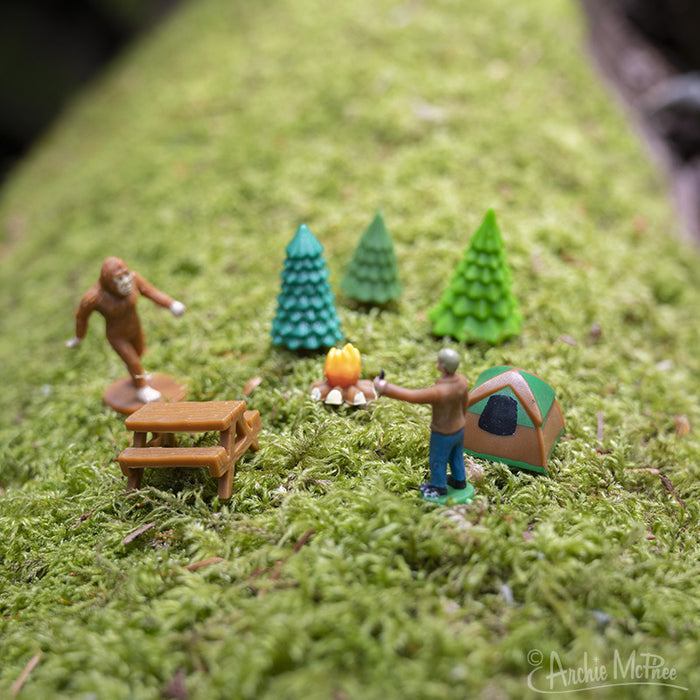 Experience a captivating miniature camping scene with the Bigfoot Basecamp, featuring soft vinyl figures set amidst tiny trees, a tent, campfire, and picnic table on a lush mossy surface—ideal for creating a distinctive desktop display.
