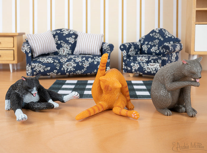 Three Cleaning Kitties figures striking yoga poses are displayed on a wooden floor, encircled by a miniature living room set with patterned sofas. Ideal for cat owners who enjoy distinctive decor.
