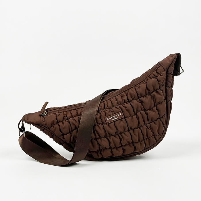 Brown crescent-shaped crossbody dog walking bag with wide strap and attached poop bag dispenser, shown on a white background.