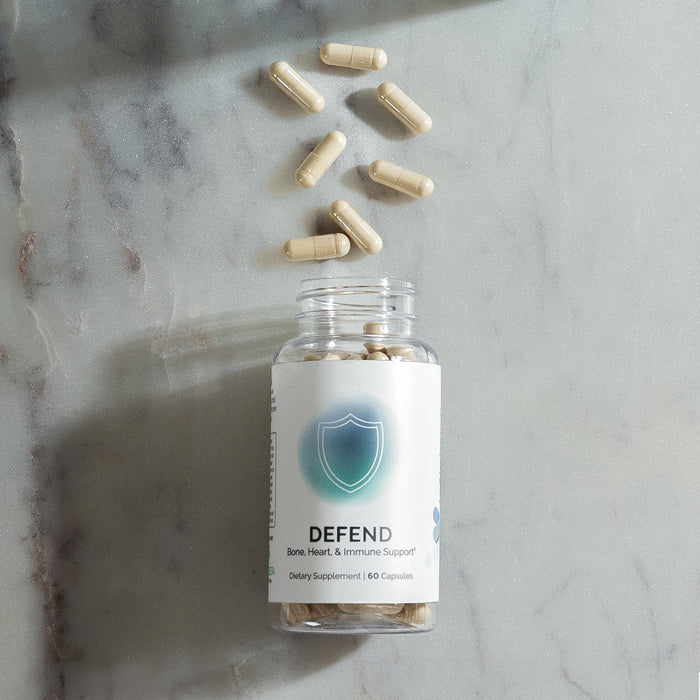 InfiniWell Defend dietary supplement bottle with open top and capsules arranged above on a marble surface.