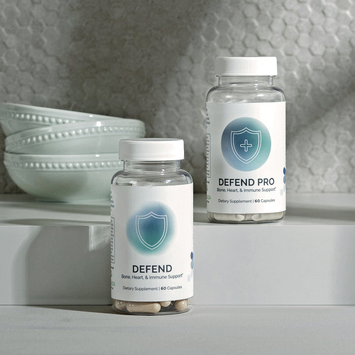 Two InfiniWell Defend dietary supplement bottles with 60 capsules displayed on a white countertop beside stacked bowls.