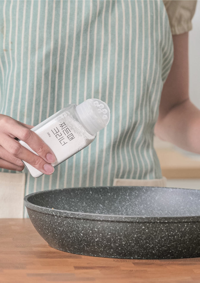 Person holding a bottle of cleaning product next to a frying pan on a wooden surface.