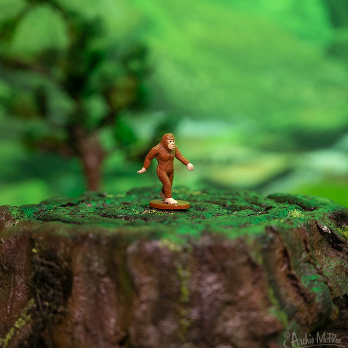 Itty Bitty Bigfoot is positioned on a mossy tree stump, enveloped by a blurred green backdrop that evokes the essence of a fantasyland forest.