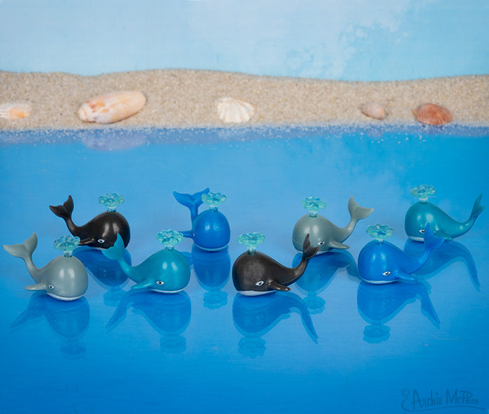 Itty Bitty Whales drifting on a blue surface with a sandy backdrop and visible shells.
