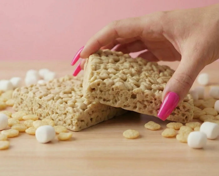 Hand with pink nails holding a marshmallow rice crispy treat on a light pink background