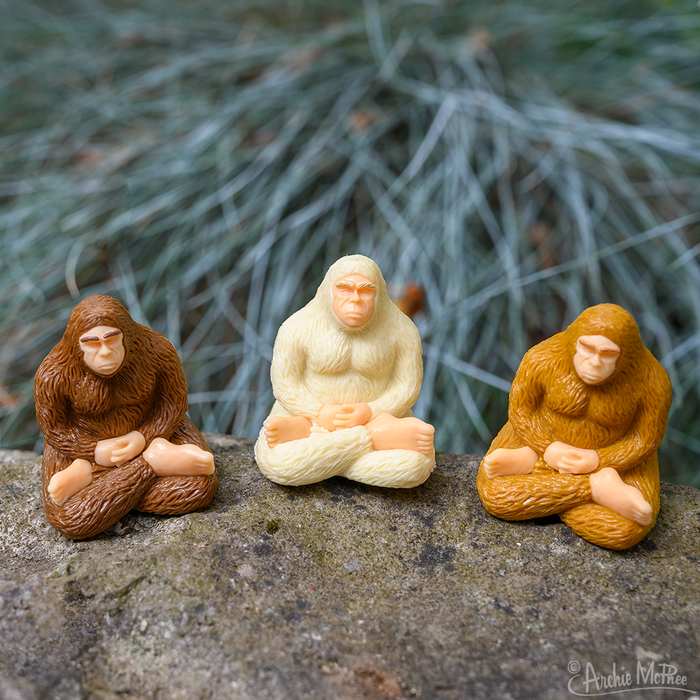 Three Meditating Bigfoot figures in brown, white, and yellow sit cross-legged on a stone surface, embodying mindfulness. Grass serves as their backdrop.
