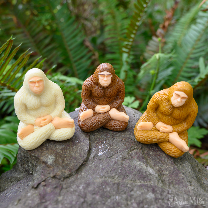 Three collectible Meditating Bigfoot figures are posed on a rock surrounded by ferns, exuding a sense of mindfulness.