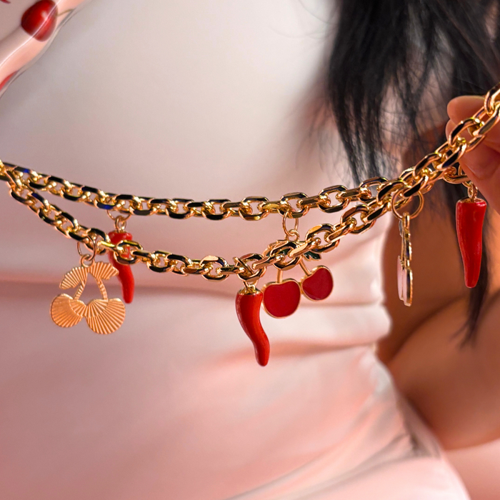 Gold chain belt with red pepper and cherry charms, held by hands wearing pink sleeves.