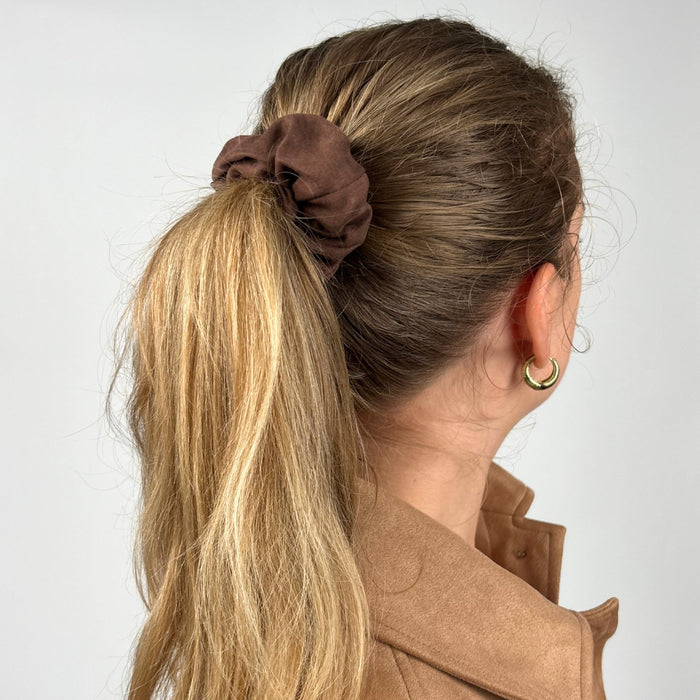 Woman wears a chocolate brown suedette scrunchie in her ponytail, gold hoop earrings, and a tan blazer.