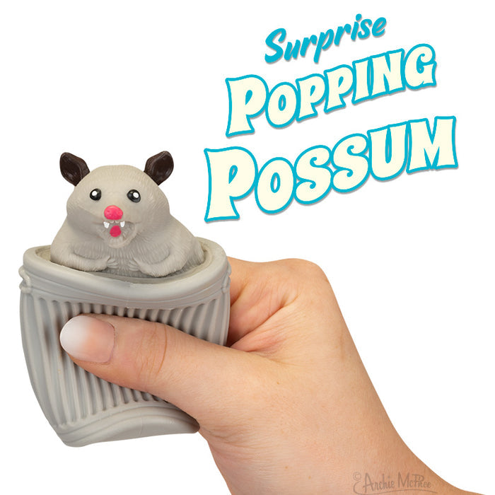 A hand squeezes the Surprise Popping Possum toy, making a possum pop out. The text reads Surprise Popping Possum. Packaged in an illustrated display box, this toy is crafted from soft rubber for endless fun and enjoyment.