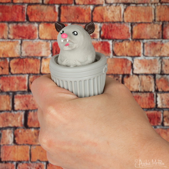 A hand holds a small, grey container with a Surprise Popping Possum toy popping out against a brick wall background. The product's name, "Surprise Popping Possum," adds charm, enhancing the playful scene while capturing attention.