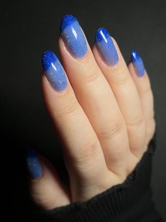 Hand with light blue to dark royal blue thermal nail polish on a dark background. Thermal nail polish with reflective glitter. Opal Theory Polish Boomerang Nebula.