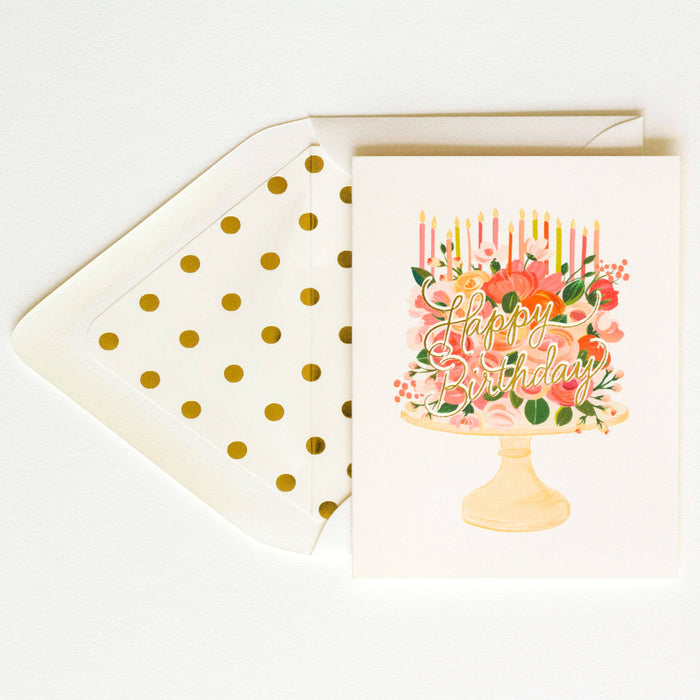 Greeting card with a birthday cake design, featuring flowers and candles, on a white background with gold dotted lines and a cream envelope.