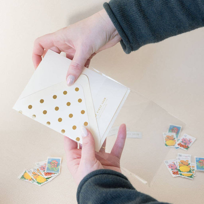 Person holding a card with gold polka dots and an envelope on a beige surface with stamps.