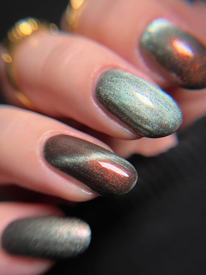 Hand with ghostly gray magnetic nail polish with orange to red shifting shimmer on a dark background. Opal Theory Polish The Pillars of Creation.