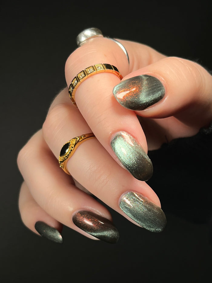 Hand with ghostly gray magnetic nail polish with orange to red shifting shimmer on a dark background. Opal Theory Polish The Pillars of Creation.