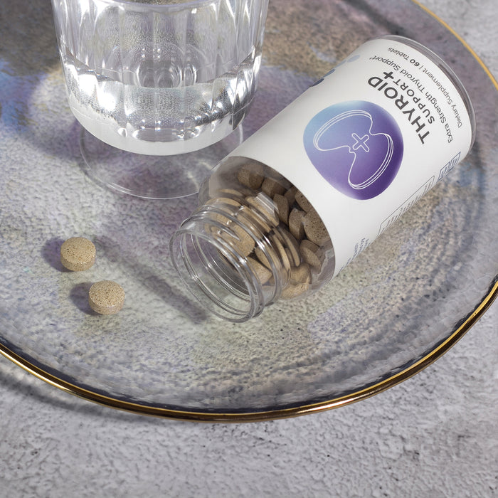 InfiniWell Thyroid Support+ Extra Strength supplement bottle with tablets spilled beside a glass of water on ceramic plate.