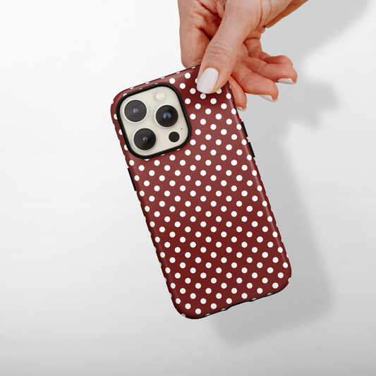 Hand holding a smartphone with a burgundy polka dot Coconut Lane tough phone case against a plain background.