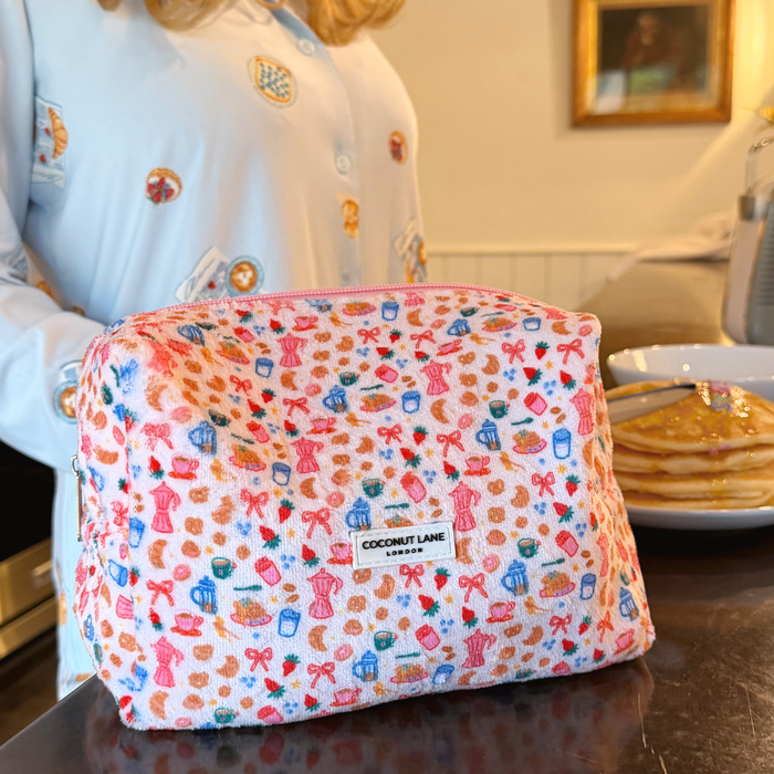 Coconut Lane towel makeup bag with Breakfast Club pattern on a counter beside pancakes and person in printed pajamas.