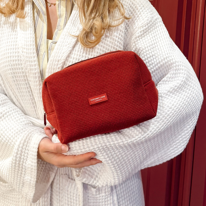 Woman in white robe holds a burgundy Coconut Lane waffle makeup bag against a red background.