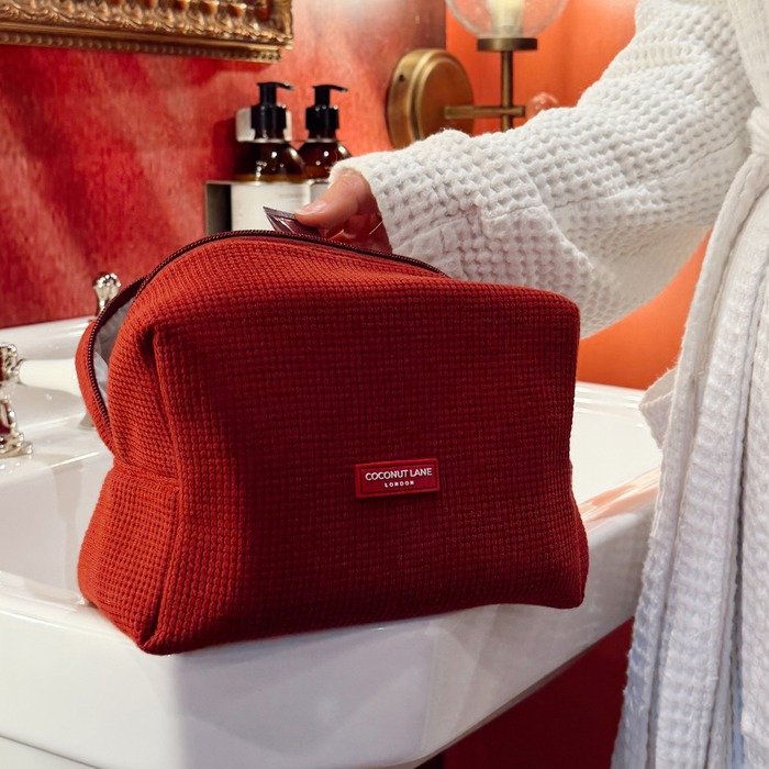 Burgundy waffle makeup bag held by a person in a white robe near a bathroom sink with soap dispensers.