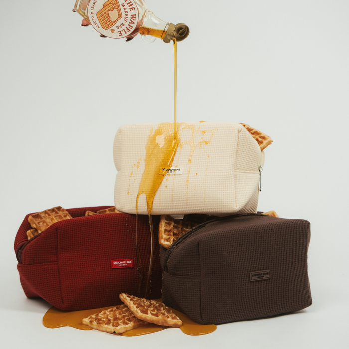 Stacked burgundy waffle makeup bags with syrup being poured on top, showing a playful, messy look.